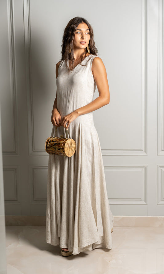 Model wears floor length dress in soft beige linen fabric with a soft pleated design that flares out from the hips down